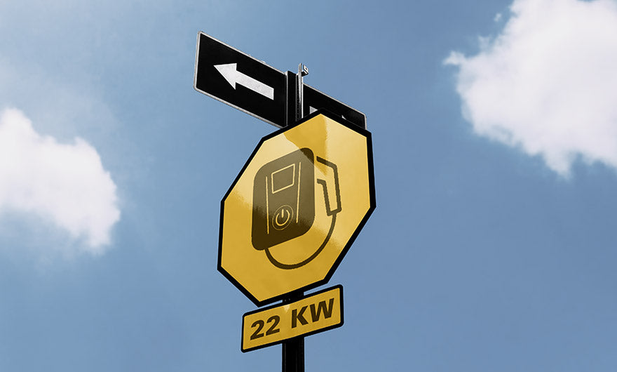 Gelbes Schild mit einem Symbol zum Laden eines Elektrofahrzeugs und darunter 22 KW; ein schwarzer Richtungspfeil zeigt nach links, vor einem blauen Himmel mit Wolken.
