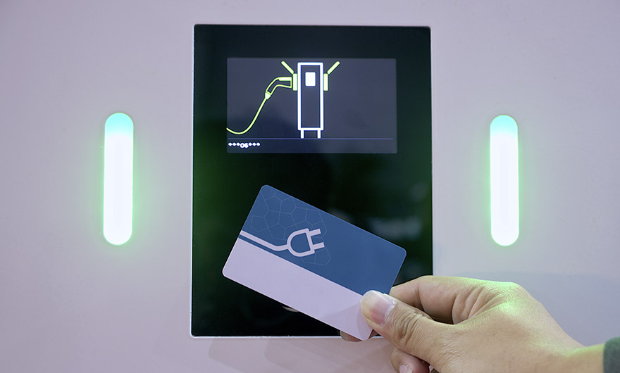 Eine Hand hält eine Karte mit einem Ladesymbol für ein Elektrofahrzeug in der Nähe eines digitalen Ladestationsbildschirms, auf dem ein Steckersymbol angezeigt wird.