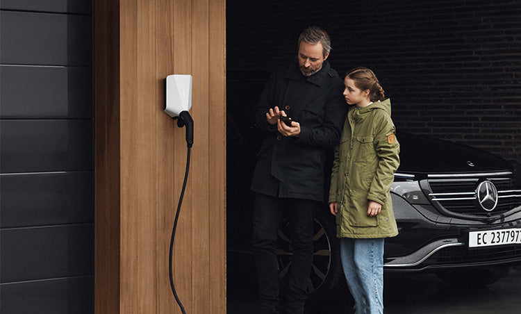 Ein Mann und ein Mädchen stehen in der Nähe einer Ladestation für Elektroautos, im Hintergrund ist ein schwarzer Mercedes-Benz geparkt. Der Mann benutzt ein Smartphone.