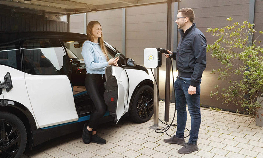 Eine Frau steht neben einem Elektroauto und hält ein Ladekabel in der Hand, während ein Mann sich darauf vorbereitet, das Kabel in eine Wandladestation zu stecken.