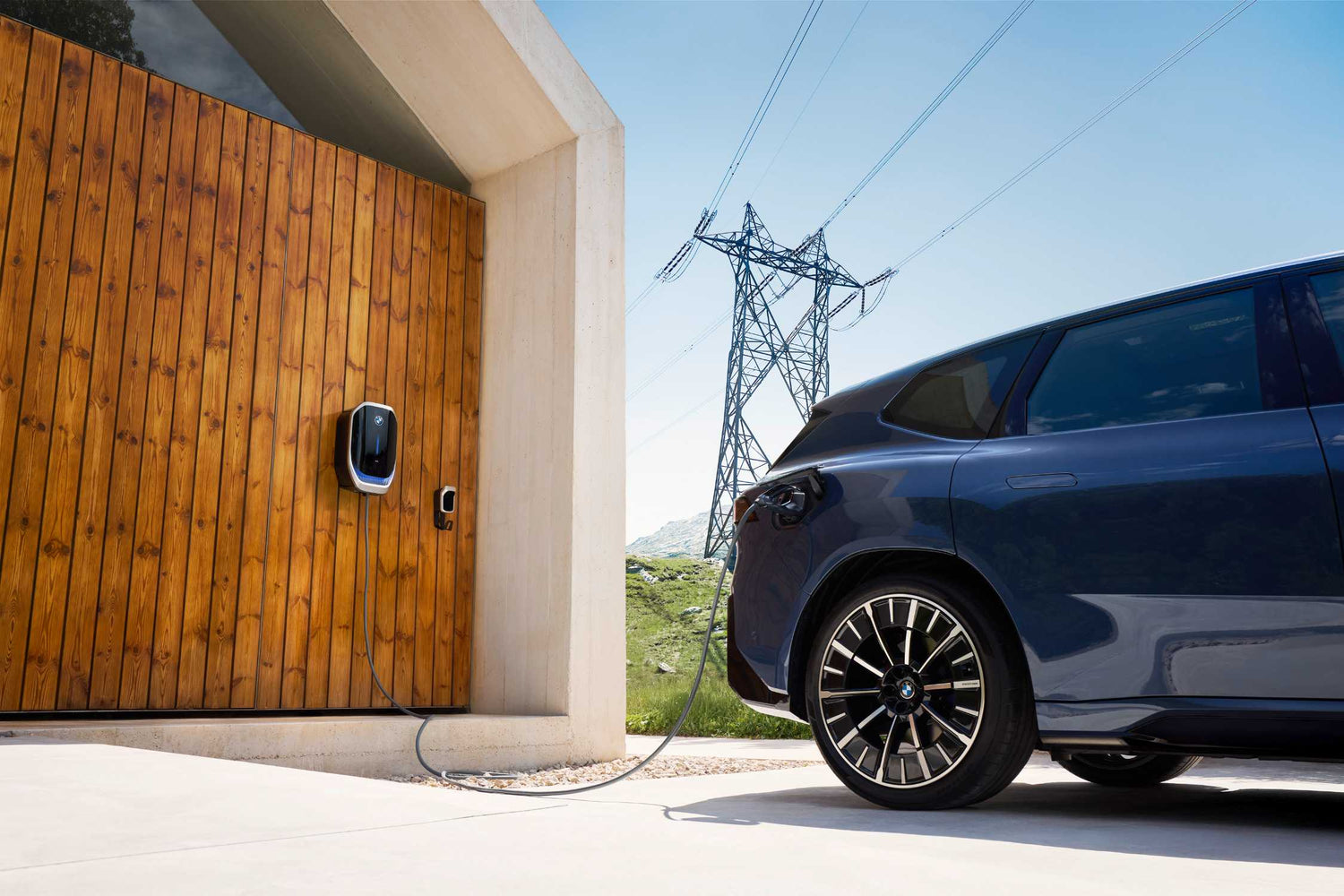 Ein dunkles Elektroauto ist an ein an der Wand montiertes Ladegerät vor einem Holzgebäude angeschlossen, mit Stromleitungen und blauem Himmel im Hintergrund.