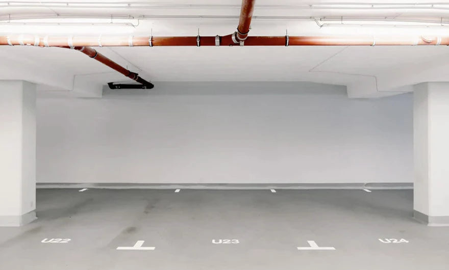 Leeres Parkhaus mit drei markierten Stellplätzen mit der Bezeichnung U22, U23 und U24, grauen Wänden und roten Rohren über dem Kopf.