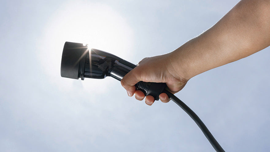 Eine Hand hält einen Ladestecker für ein Elektrofahrzeug in den hellen Himmel, wobei das Sonnenlicht durch den Stecker scheint.