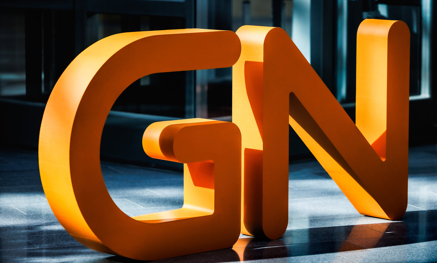 Große, orangefarbene, dreidimensionale Buchstaben GN stehen auf einem polierten Boden in einem Innenraum, der von natürlichem Licht beleuchtet wird.