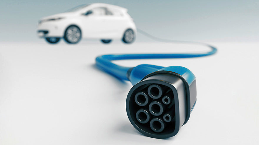 Nahaufnahme eines Ladekabels für Elektrofahrzeuge im Vordergrund, mit einem unscharfen weißen Elektroauto im Hintergrund.