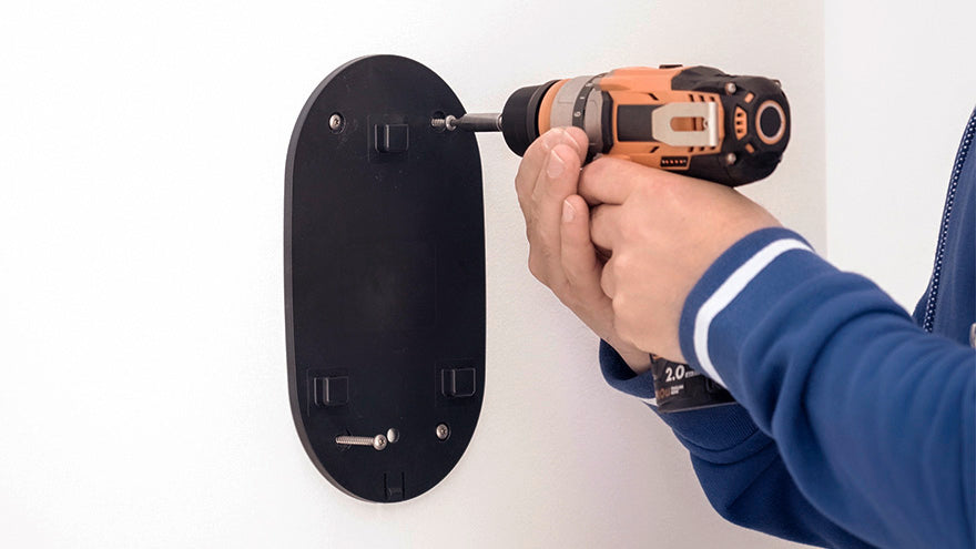 Eine Person verwendet einen Akkubohrer, um eine schwarze Montageplatte an einer weißen Wand zu befestigen.