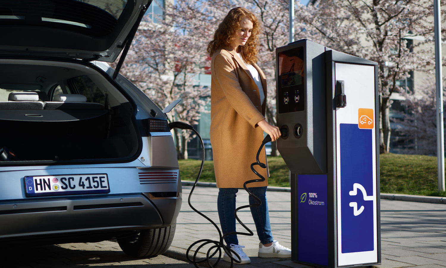 Eine Frau steht neben einem Elektroauto und steckt an einem sonnigen Tag ein Ladekabel in eine Ladestation.