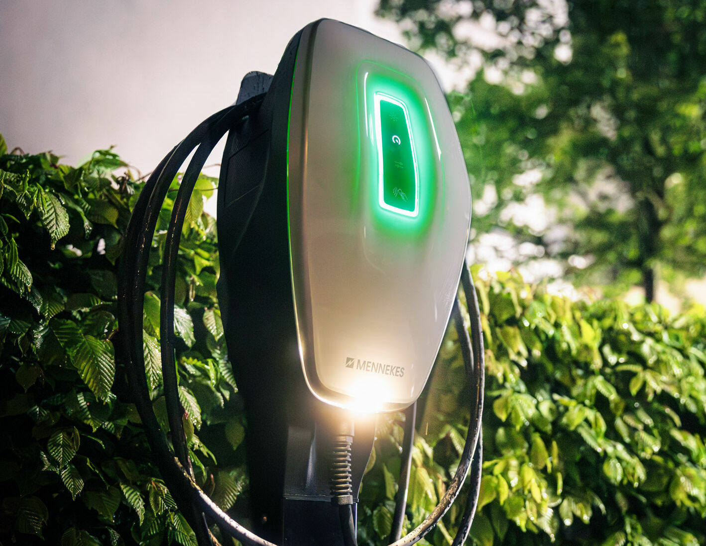 Eine wandmontierte Mennekes AMTRON® 4You 510 C2 Ladestation von Mennekes mit leuchtend grünem Licht ist im Außenbereich vor einer Hecke installiert.