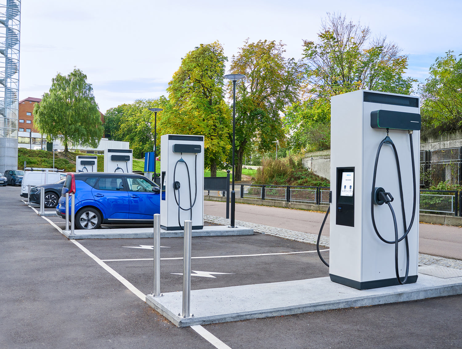 Mehrere Ladestationen für Elektrofahrzeuge mit geparkten und eingesteckten Autos auf einem Außenparkplatz mit Bäumen und Gebäuden im Hintergrund.