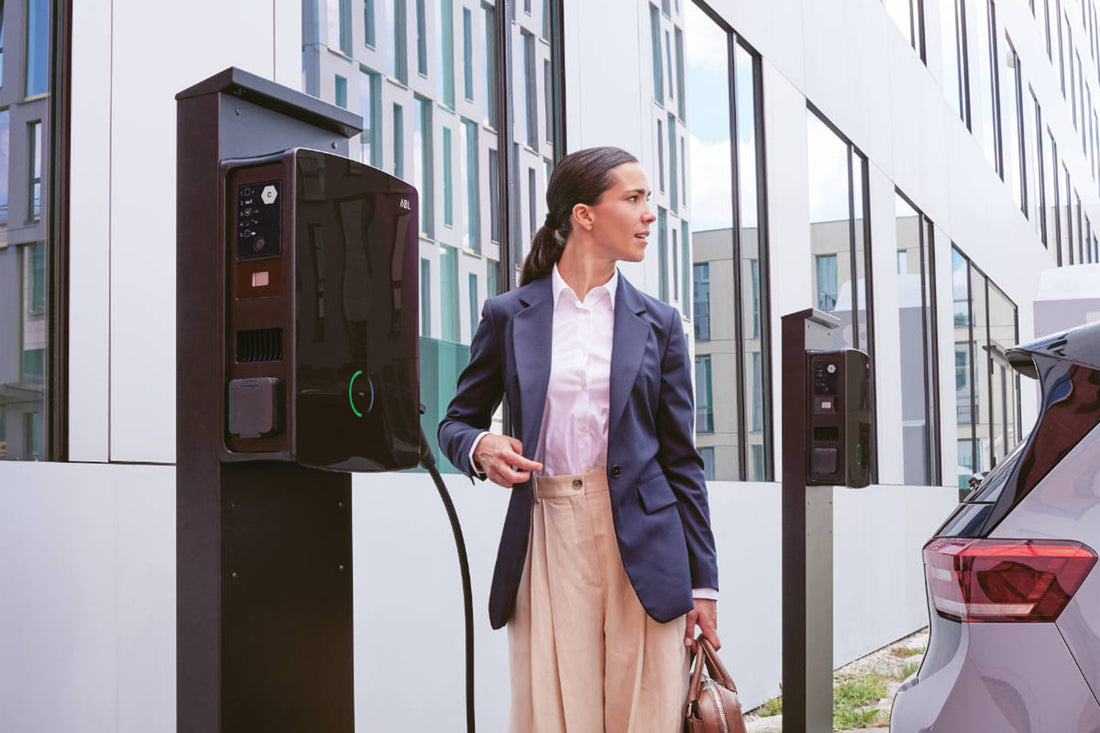 Eine Frau in Businesskleidung steht neben einer ABL eM4-Ladestation und hält ein Kabel in der Nähe eines geparkten Autos vor einem modernen Bürogebäude – und präsentiert die preisgekrönte Marke ABL, die mit dem Design Plus Award 2022 ausgezeichnet wurde.