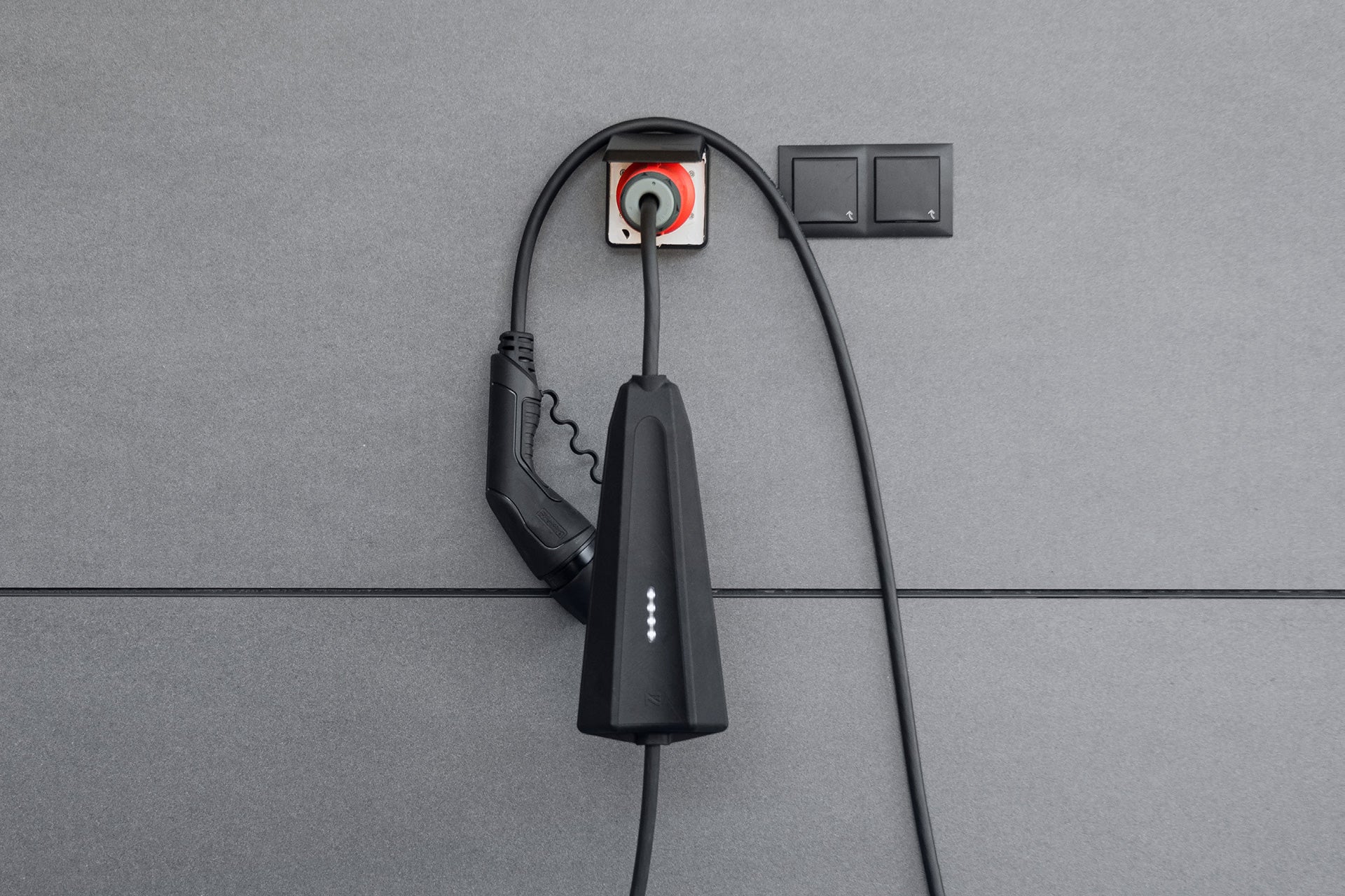 Ein NEcharge One-Ladekabel von NEcharge ist an eine Steckdose an einer grau gefliesten Wand angeschlossen und hebt die Funktionen seiner tragbaren Ladestation hervor.