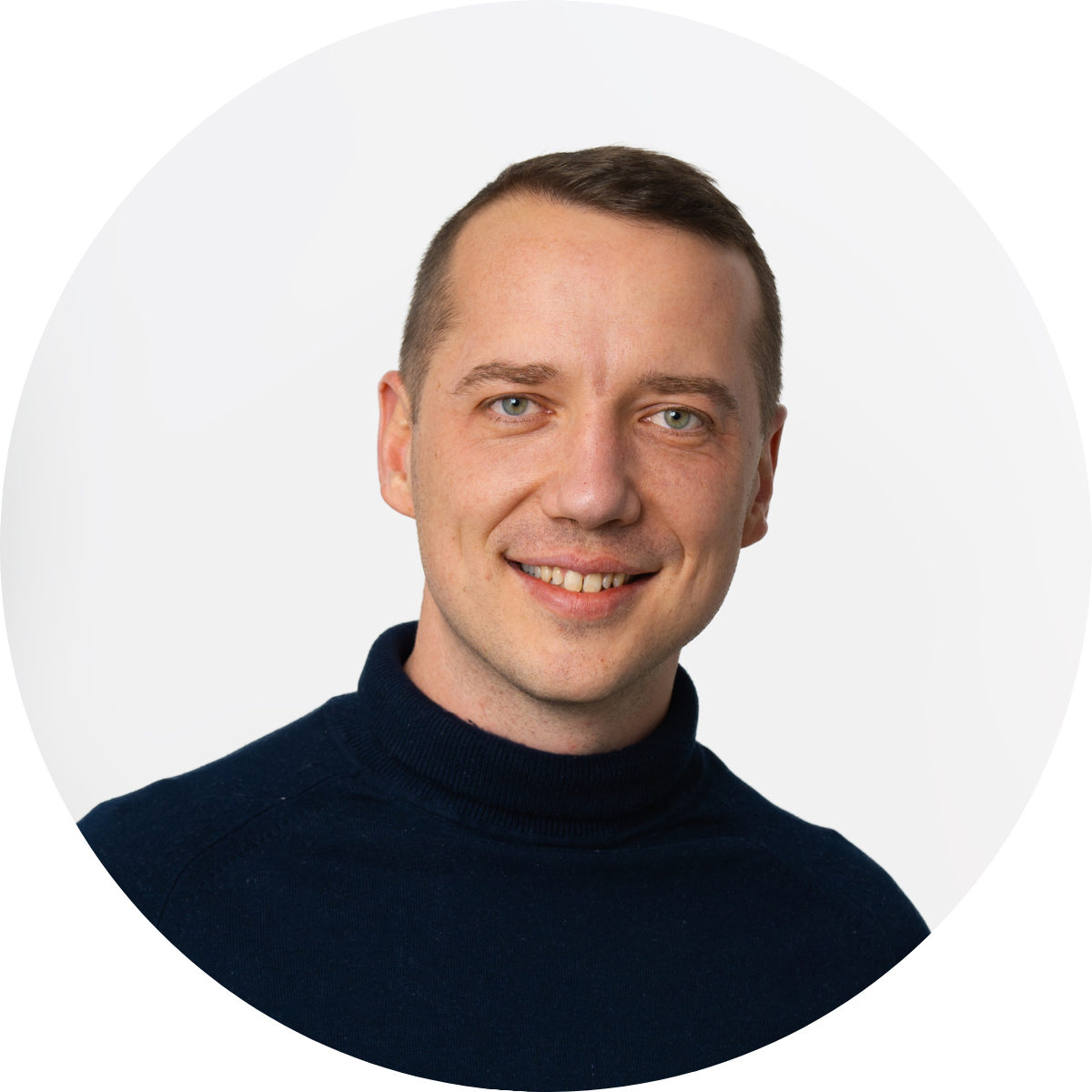 Ein Mann mit kurzem hellbraunem Haar und einem dunklen Rollkragenpullover lächelt vor einem schlichten weißen Hintergrund in die Kamera.