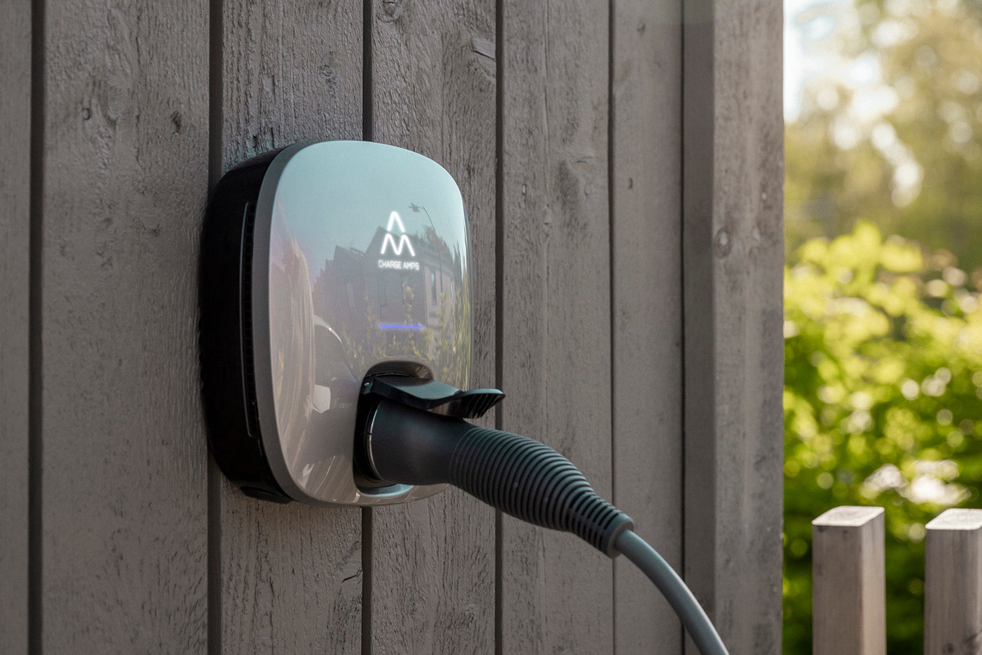 Eine wandmontierte Ladestation für Elektrofahrzeuge Charge Amps Luna von Charge Amps ist an einem Holzzaun befestigt, das Ladekabel ist eingesteckt, im Hintergrund ist Grün zu sehen.