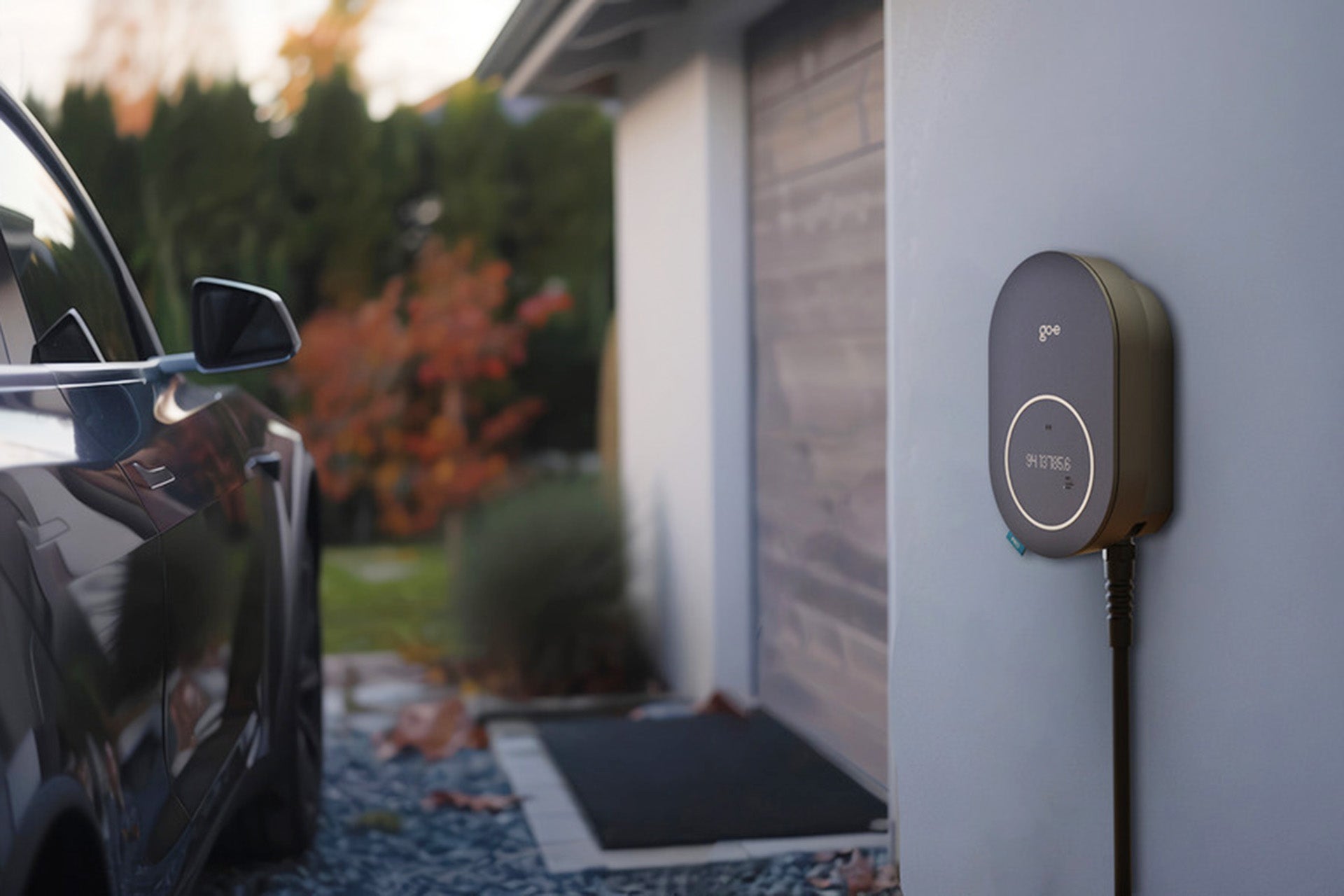 Das go-e Charger PRO CABLE von go-e ist an einer Außenwand montiert, Plug&Charge-fähig, mit einem in der Nähe geparkten Auto und einem geschlossenen Garagentor im Hintergrund.