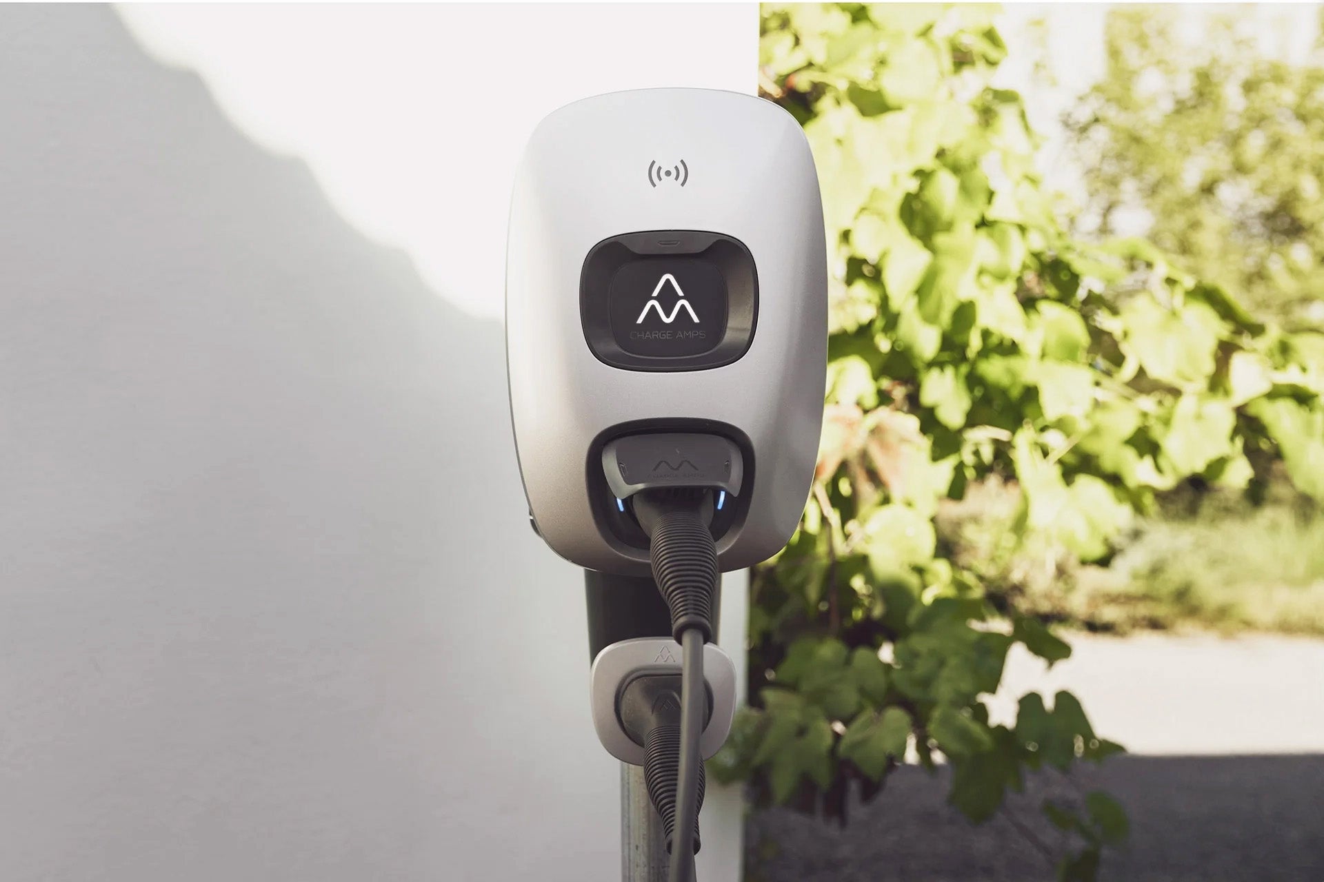 Eine Nahaufnahme der Charge Amps Dawn 22 kW Typ 2 AC-Ladestation von Charge Amps, montiert an einer weißen Wand mit Energieeffizienzfunktionen, vor einem Hintergrund aus Grün und Sonnenlicht.