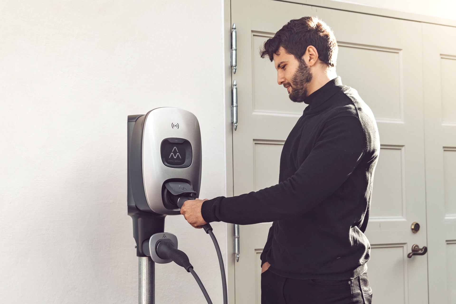 Ein Mann im schwarzen Pullover steckt ein Ladekabel in sein Elektrofahrzeug und nutzt dafür den Charge Amps Dawn, ein wandmontiertes 22-kW-Typ-2-AC-Ladegerät von Charge Amps, um die Energieeffizienz zu optimieren.