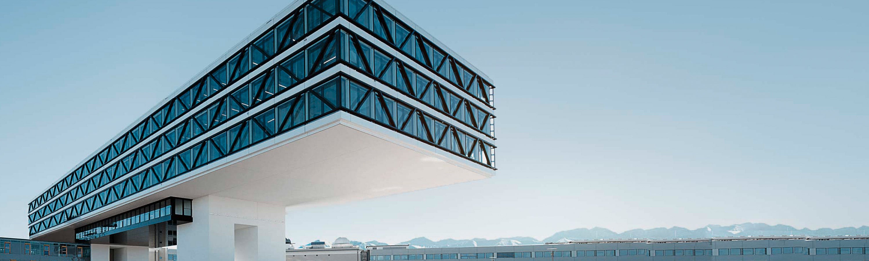 Ein modernes Gebäude mit geometrischen Obergeschossen aus Glas und Stahl erstreckt sich horizontal über einer weißen Stützstruktur vor einem klaren Himmel.