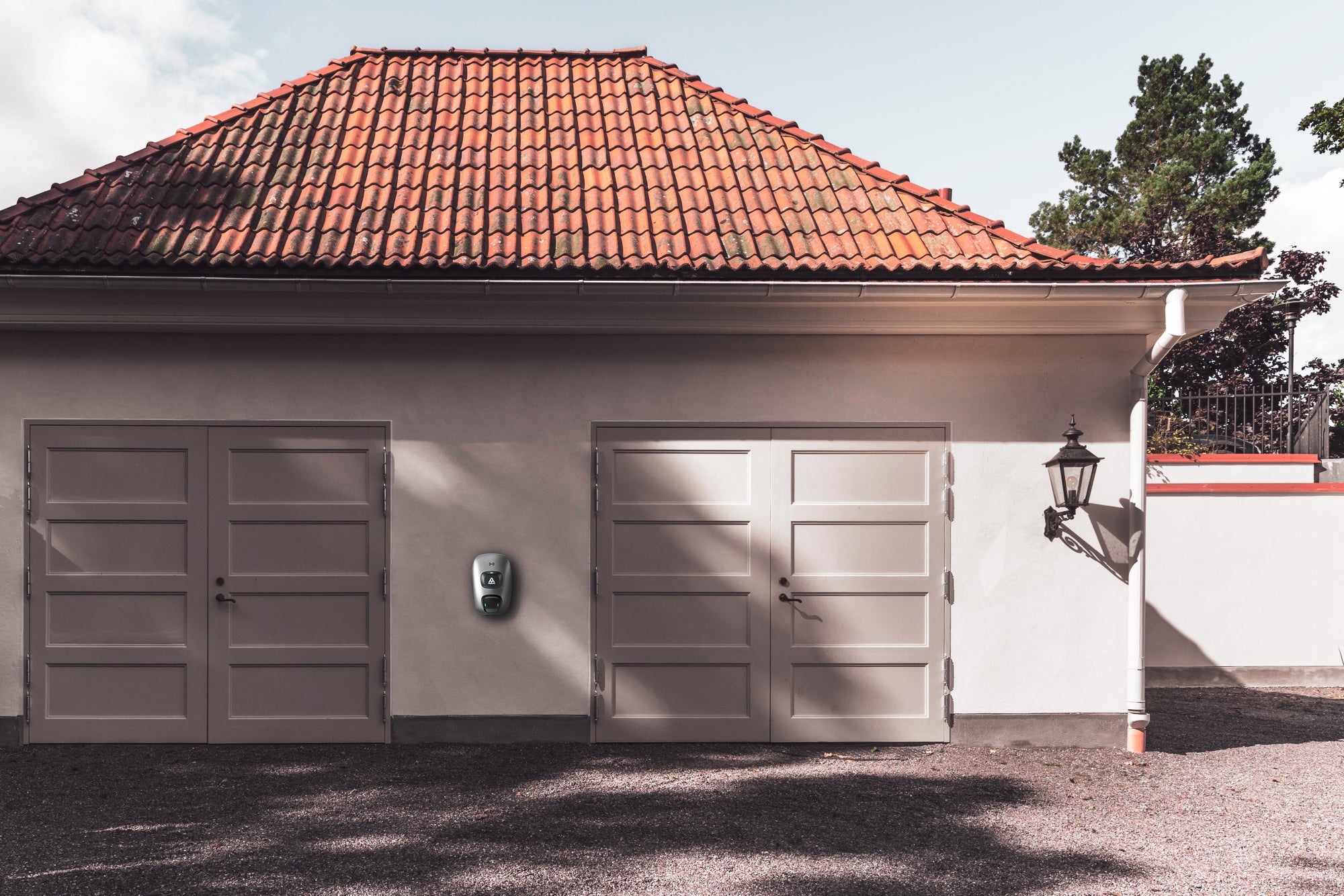 Ein Gebäude mit zwei geschlossenen Garagentoren, einem dazwischen montierten Charge Amps Dawn 22-kW-Typ-2-AC-Ladegerät von Charge Amps, einem Ziegeldach und einer Außenlampe an der rechten Wand.