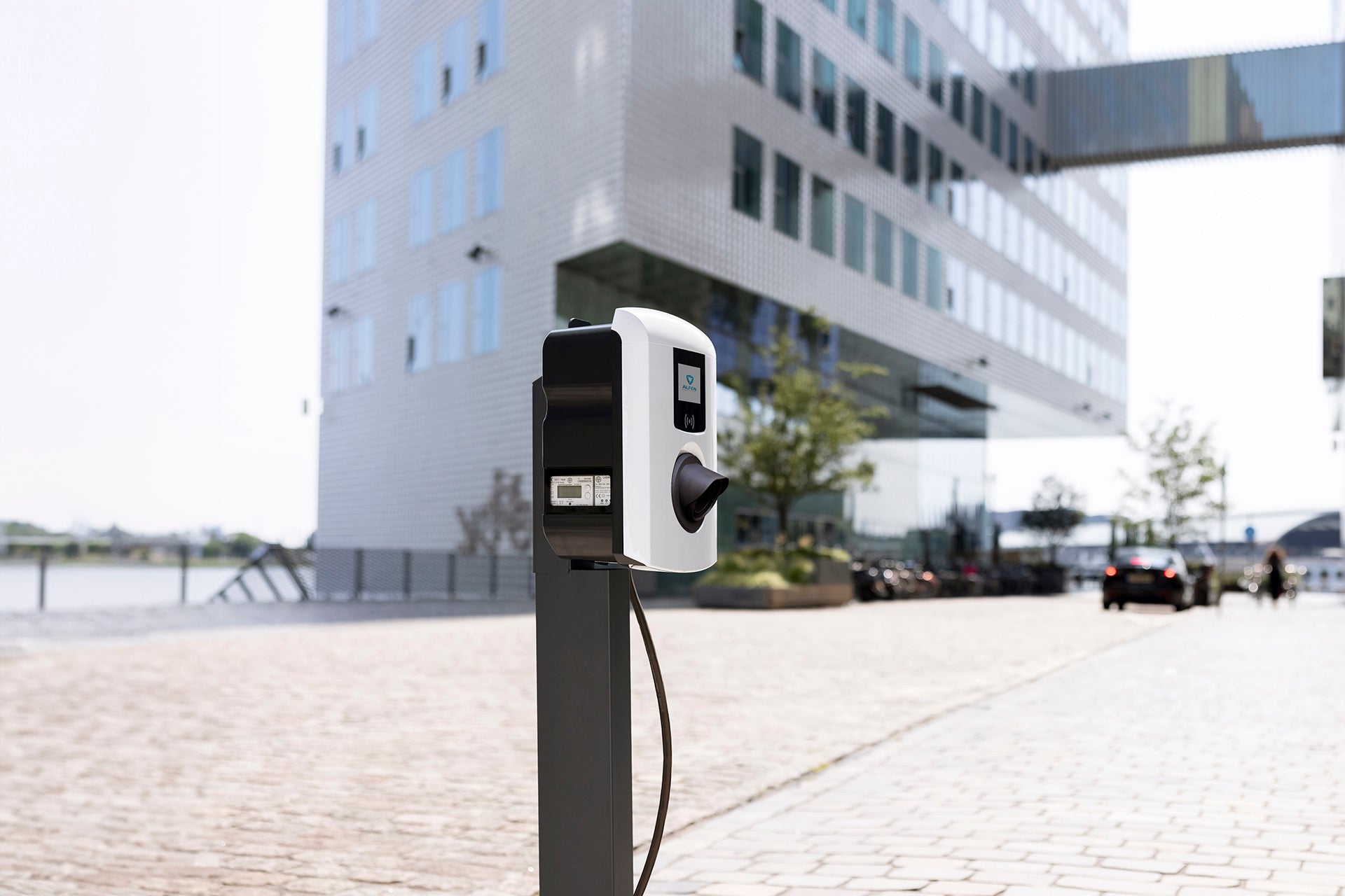 Eine benutzerfreundliche Alfen Eve Single Pro-Line Ladestation von Alfen steht auf dem Kopfsteinpflaster vor einem modernen Glas- und Stahlbürogebäude, mit Autos und Bäumen im Hintergrund.