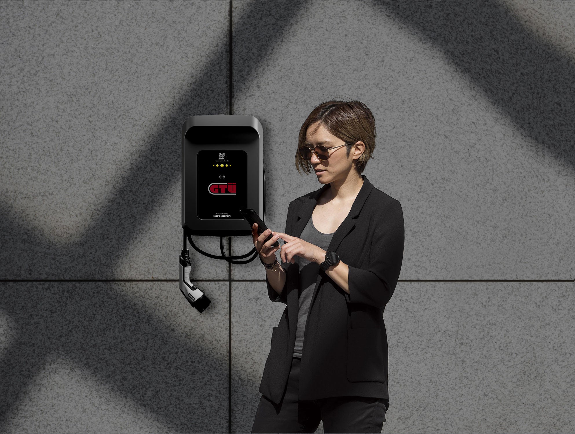 Eine Person mit Sonnenbrille und dunkler Kleidung steht neben einer GTÜ Wallbox WL60 von GTÜ und nutzt ein Smartphone mit RFID-Zugang vor einer grau gefliesten Wand.