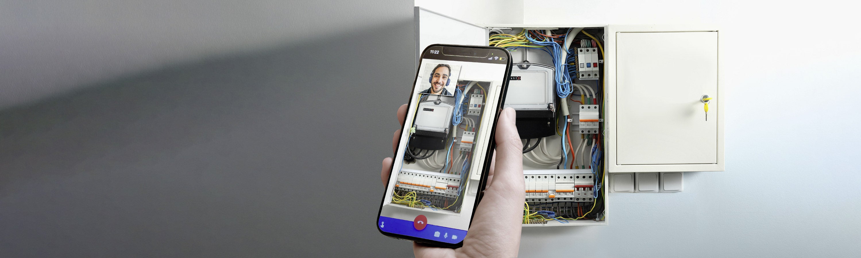 Eine Person hält ein Smartphone, auf dem ein Videoanruf läuft, vor einem offenen Schaltschrank mit sichtbaren Kabeln und Komponenten.