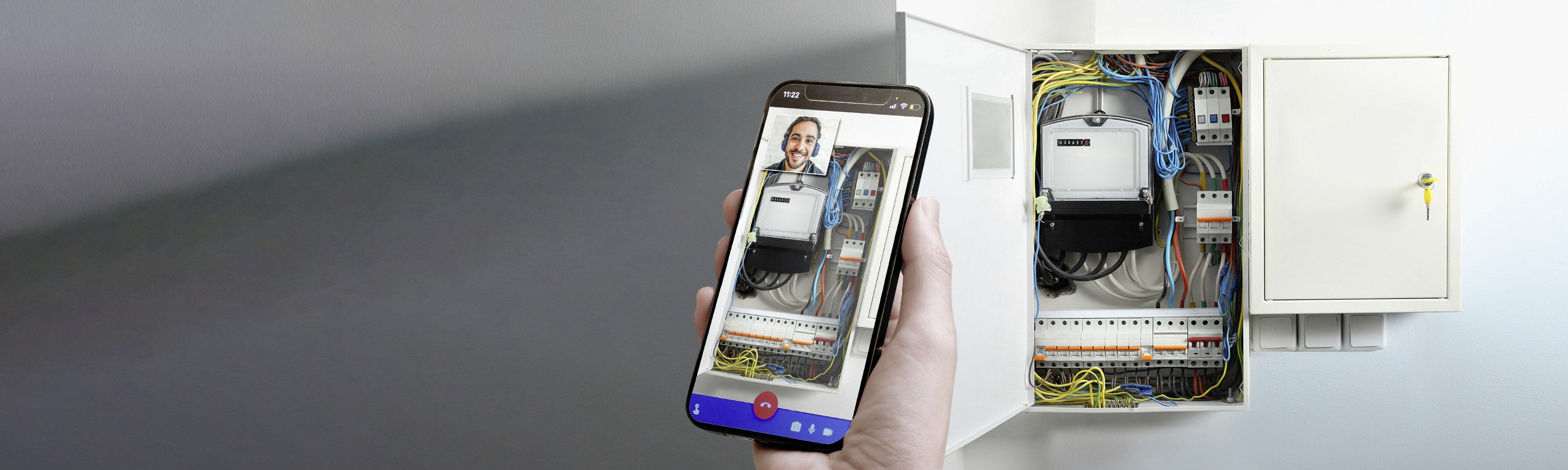 Eine Hand hält ein Smartphone, auf dem ein Videoanruf läuft, vor einem offenen Schaltschrank mit verschiedenen Kabeln und Komponenten.