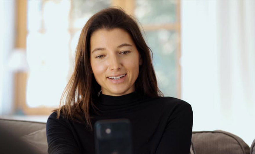 Frau mit langen braunen Haaren in schwarzem Rollkragenpullover sitzt auf einem Sofa, lächelt in ihr Smartphone und liest über den e-mobilio Video-Installations-Check vom e-mobilio Shop, im Hintergrund ein Fenster.