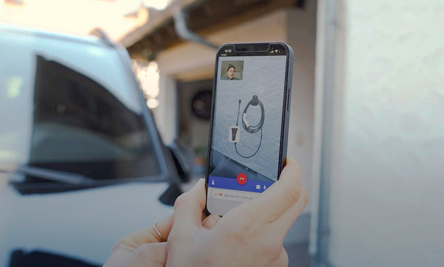 Eine Person hält ein Smartphone, auf dem ein Videoanruf läuft; auf dem Bildschirm ist ein aufgerolltes Ladekabel für eine Wallbox-Elektroinstallation zu sehen, hervorgehoben wird der DVS Video-Installations-Check von e-mobilio Shop.