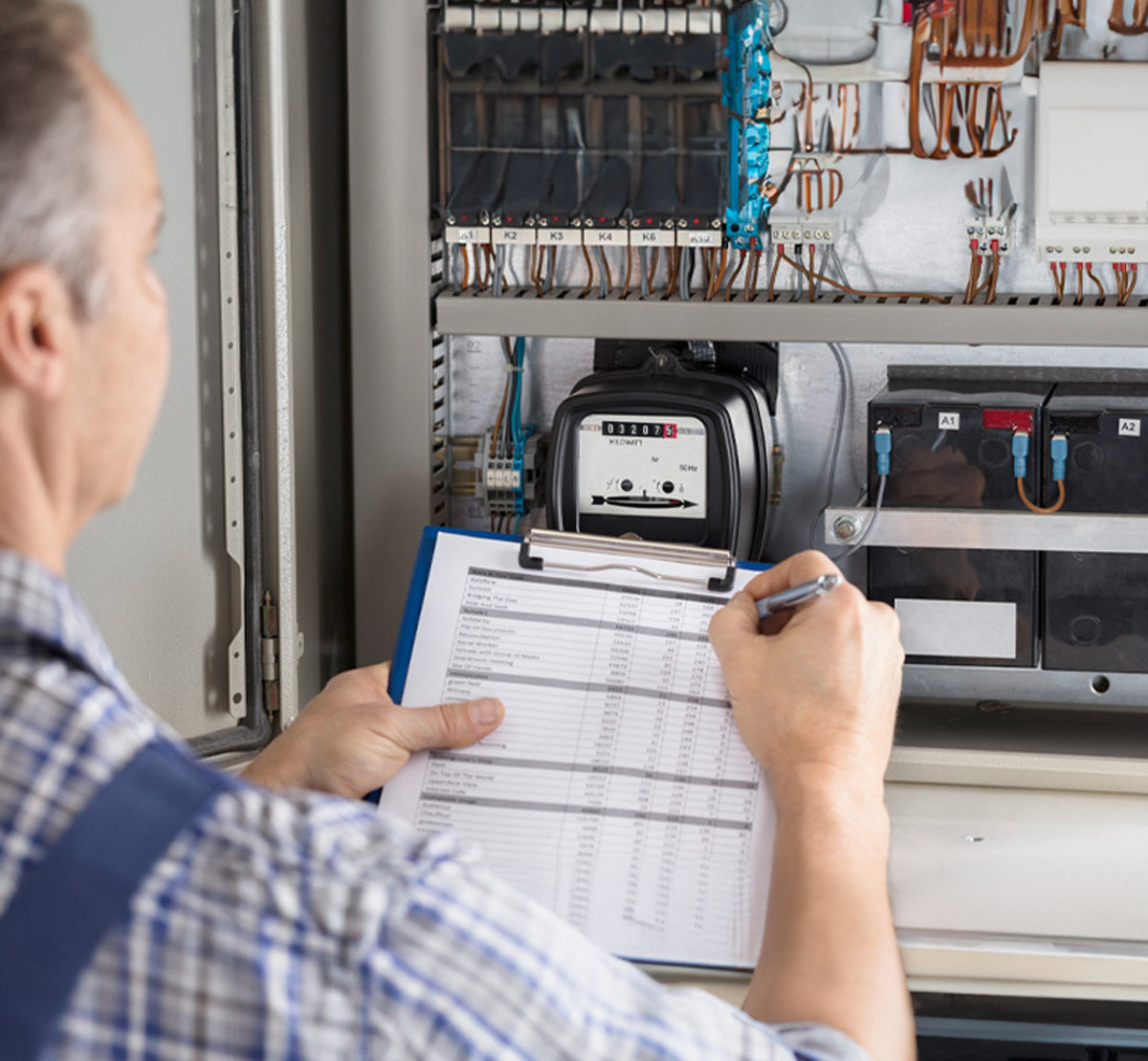Eine Person prüft und erfasst Daten von einem offenen elektrischen Bedienfeld und hält dabei ein Klemmbrett mit einer ausgedruckten Checkliste oder einem Protokollblatt in der Hand.