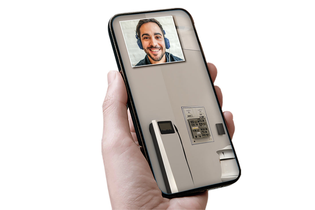 Eine Hand hält ein Smartphone, auf dem ein Videoanruf mit einem lächelnden Mann und der e-mobilio Video-Installations-Check vom e-mobilio Shop zu sehen sind, auf dem ein Bild einer Wallbox-Elektroinstallation oder eines Zahlungsterminals zu sehen ist.