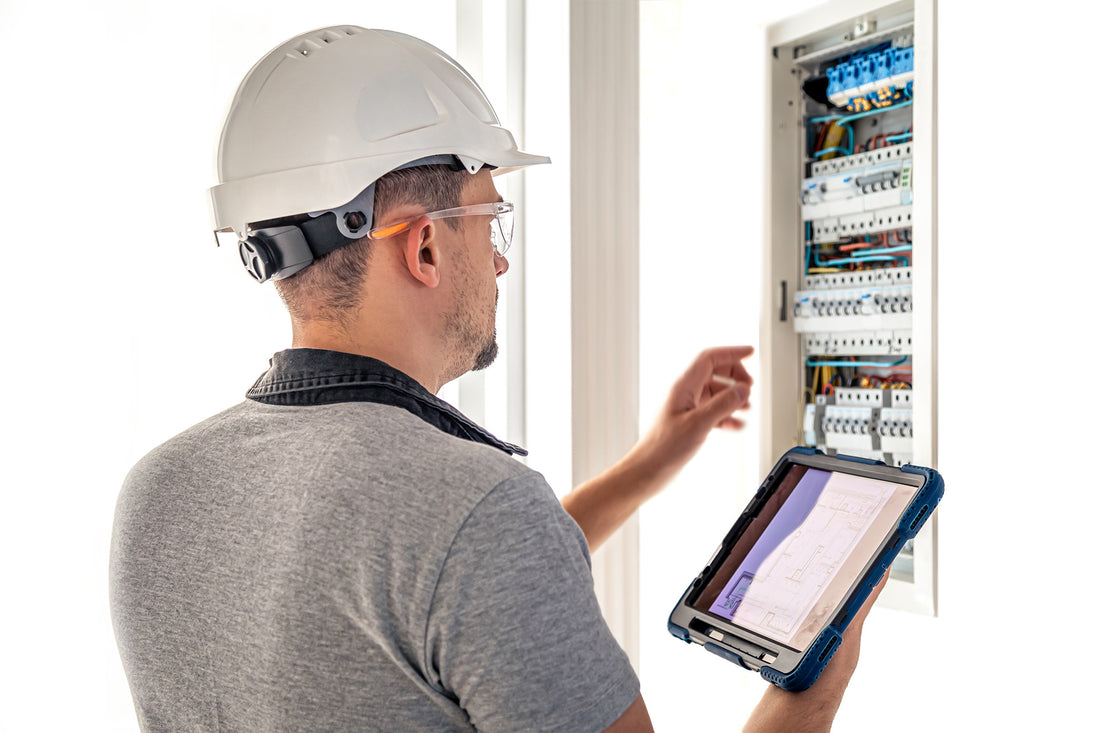 Eine Person mit Schutzhelm und Schutzbrille inspiziert einen offenen Schaltschrank und hält ein Tablet in der Hand, auf dem die Ladepunktkonfiguration aus dem e-mobilio Shop angezeigt wird.
