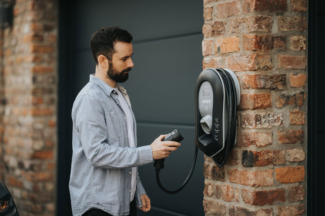 Ein Mann hält ein Ladekabel in der Hand und ist bereit, es an ein an einer Ziegelwand montiertes 22-kW-Ladegerät vom Typ myenergi zappi V2.1 V2H22 anzuschließen – perfekt für den Einsatz mit Photovoltaiksystemen und mit flexiblen Lademodi.
