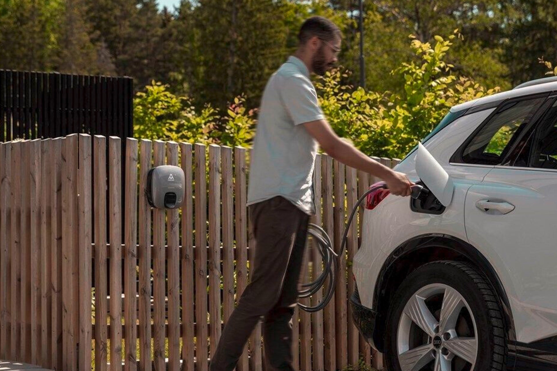 Ein Mann schließt ein Ladekabel an ein weißes Elektrofahrzeug an, das an einem Holzzaun geparkt ist. Dazu verwendet er die Ladestation Charge Amps Luna von Charge Amps, die mit zukunftssicherer Technologie und innovativem Lastausgleich ausgestattet ist.