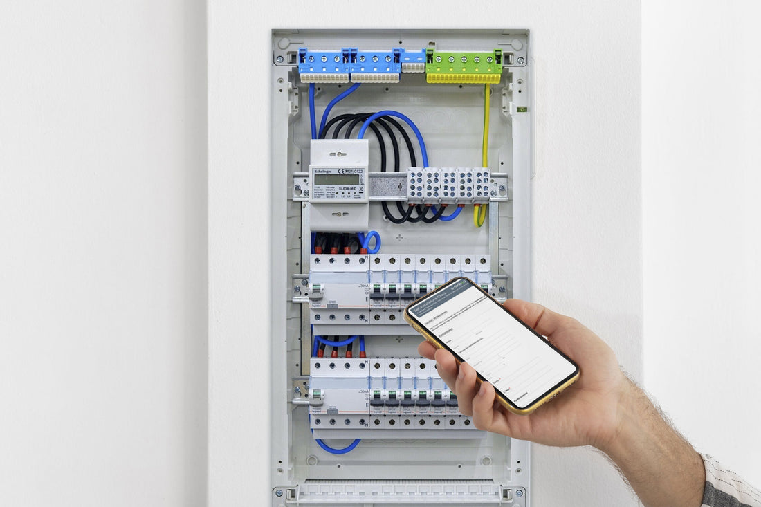 Jemand hält ein Smartphone mit der App „e-mobilio Self-Installation-Check“ des e-mobilio Shops vor einen offenen Schaltschrank mit sichtbaren Leitungen und Sicherungsautomaten.