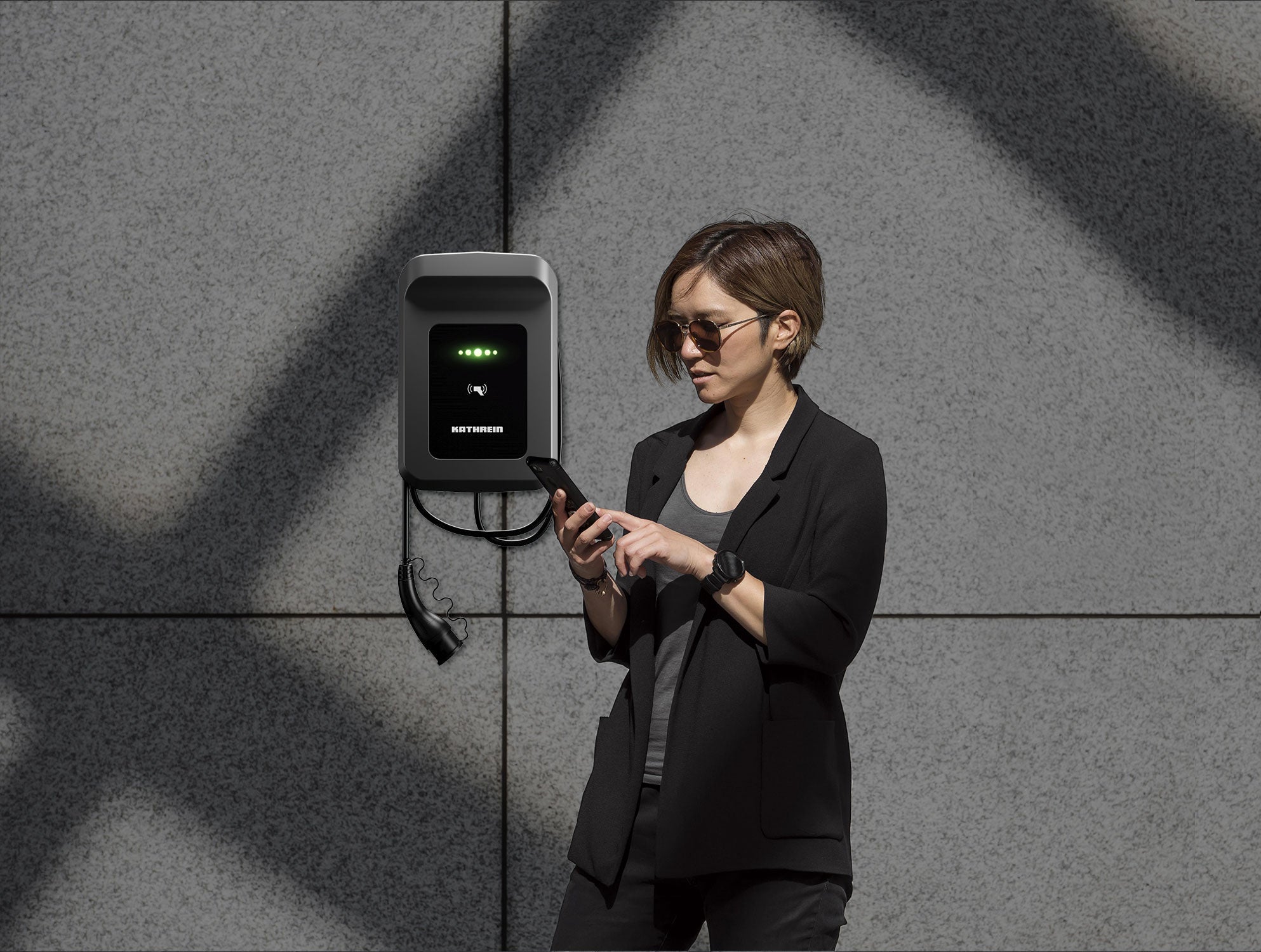 Eine Person mit Sonnenbrille nutzt ein Smartphone neben einer wandmontierten Kathrein KWB-AC60E-Ladestation für Elektrofahrzeuge mit RFID-Autorisierung.