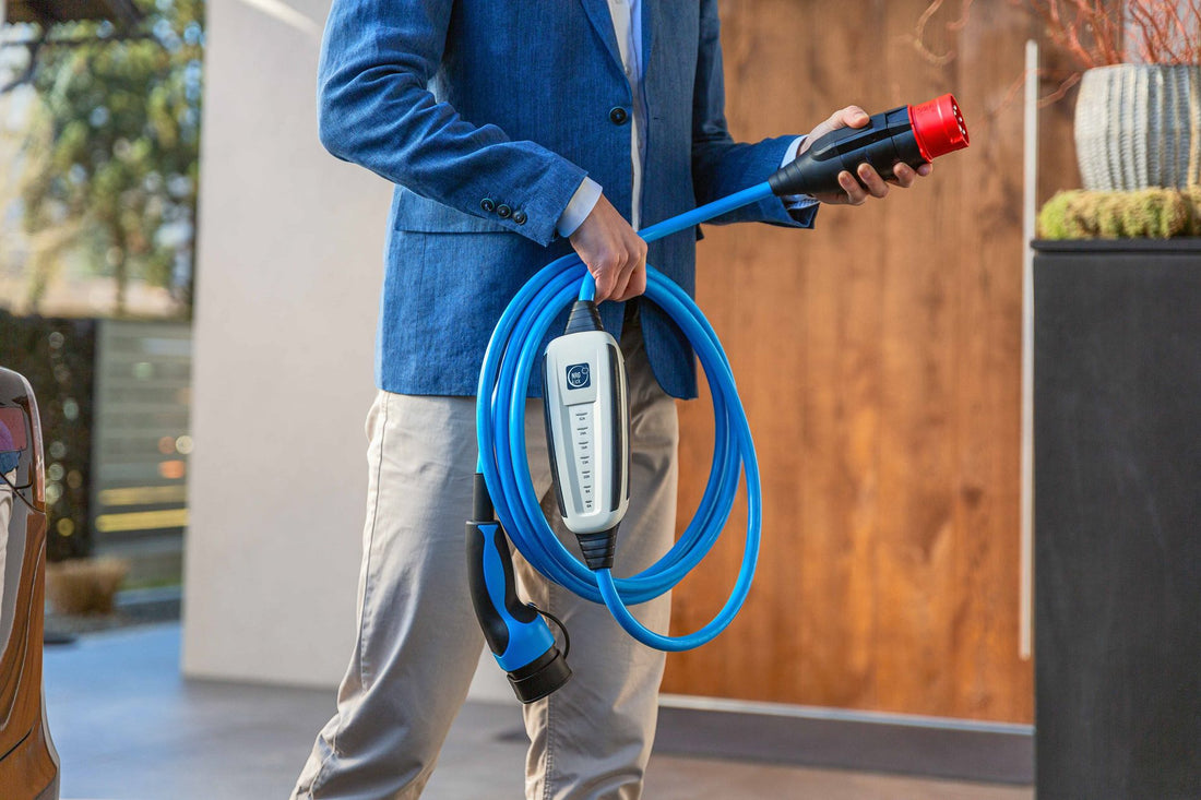 Eine Person in einem blauen Blazer hält einen Typ-2-Stecker und ein blaues EV-Ladekabel, steht im Freien neben einem modernen Gebäude und präsentiert das NRGkick-Set von NRGkick, eine mobile Ladeeinheit mit 22 kW Ladeleistung.