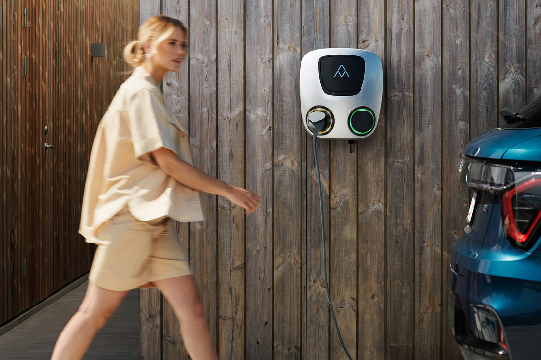 Eine Frau geht an einer Ladestation für Elektrofahrzeuge Charge Amps Aura von Charge Amps mit RFID-Autorisierung vorbei, die an einer Holzwand montiert ist, während rechts teilweise ein Auto zu sehen ist.