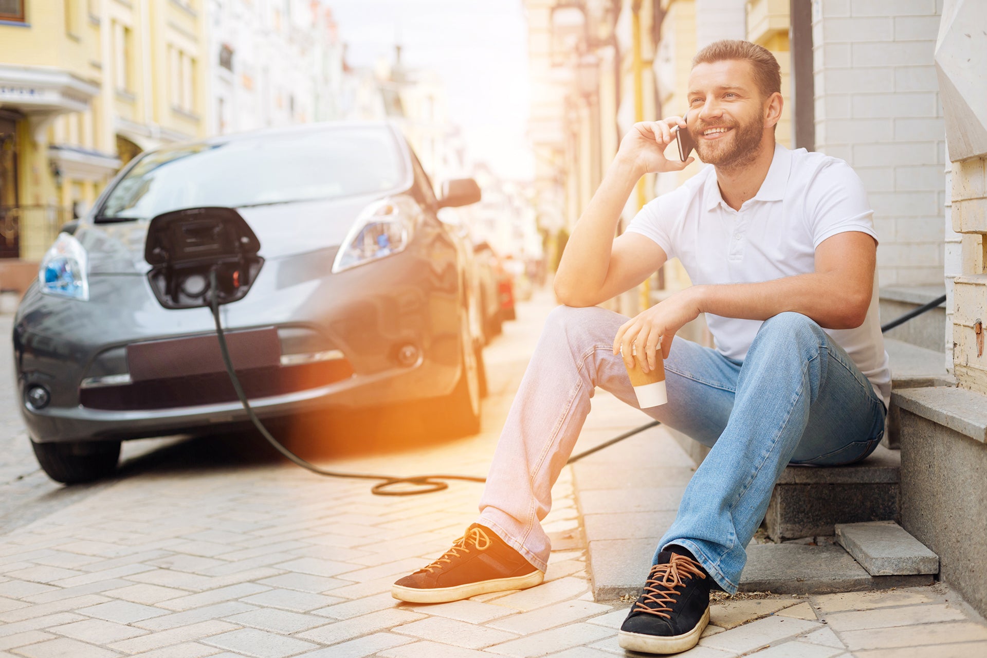 Ein Mann sitzt auf einer Außentreppe, hält eine Tasse in der Hand und telefoniert, während sein Elektroauto in der Nähe mit dem Ladekabel Typ 2 von LAPP aufgeladen wird.