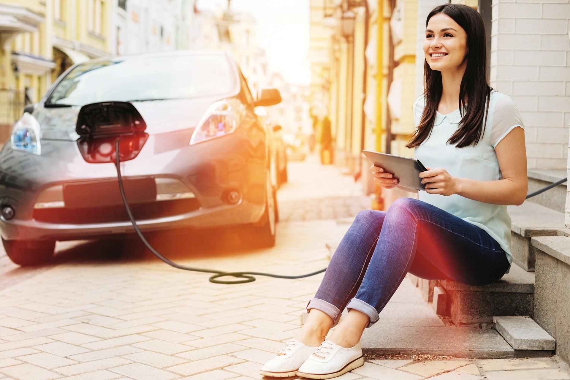 Eine Frau sitzt lächelnd mit einem Tablet auf einer Außentreppe, während ihr Elektroauto in der Nähe mit einem LAPP Ladekabel Typ 2 von LAPP auf einer Stadtstraße aufgeladen wird.
