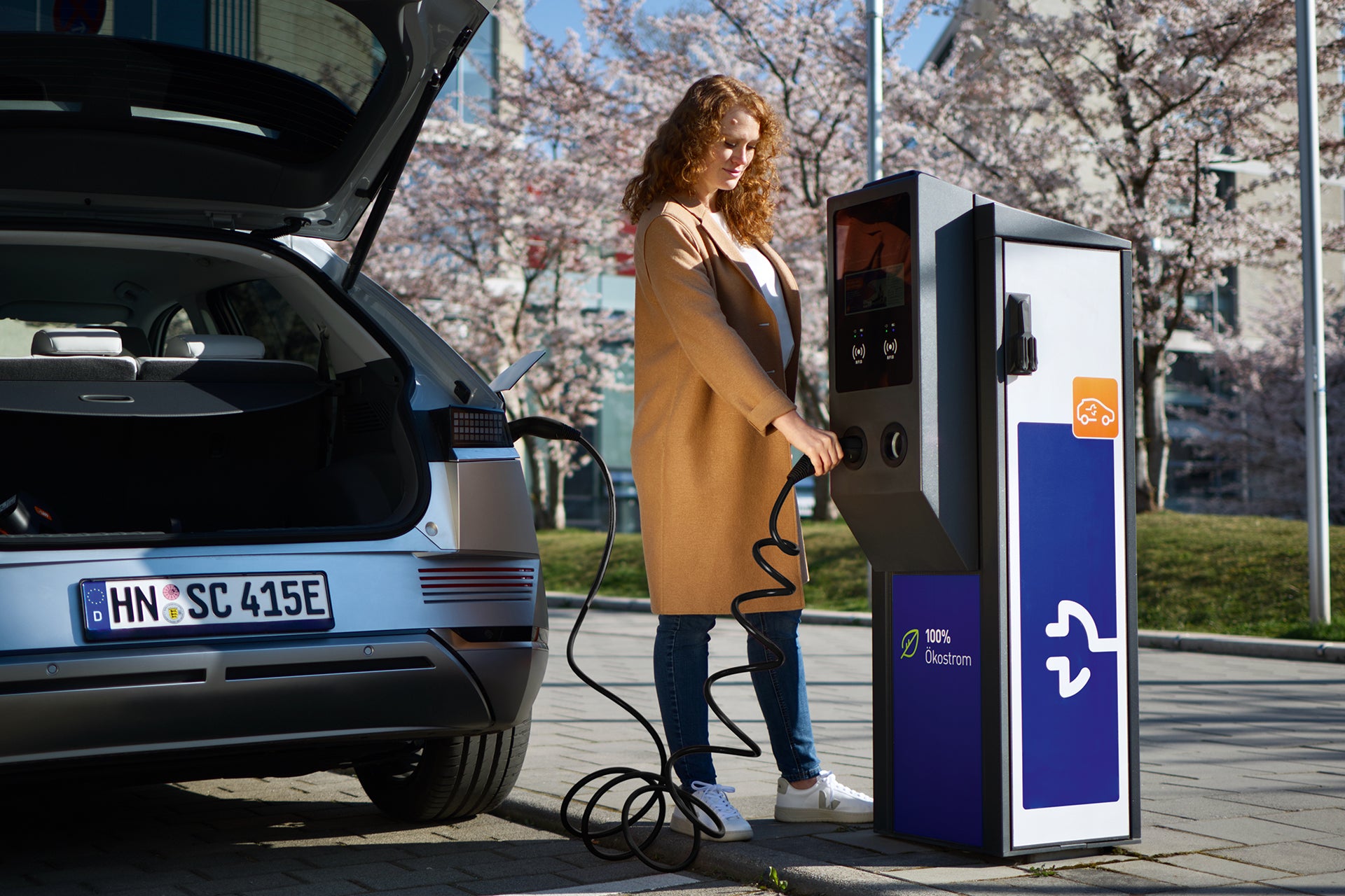 Eine Frau in einem hellbraunen Mantel schließt an einer öffentlichen Ladestation ein Lapp-Helix-Ladekabel Typ 2 von LAPP an ein Elektroauto an und demonstriert so, wie praktisch es ist, Elektroautos an einem sonnigen Tag aufzuladen.
