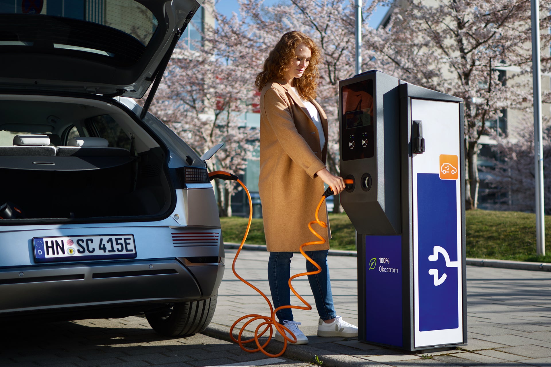 Eine Frau lädt ein Elektroauto im Freien an einer öffentlichen Ladestation mit dem LAPP Helix Ladekabel Typ 2 auf. Der Kofferraum ist geöffnet, im Hintergrund sind Bäume zu sehen.