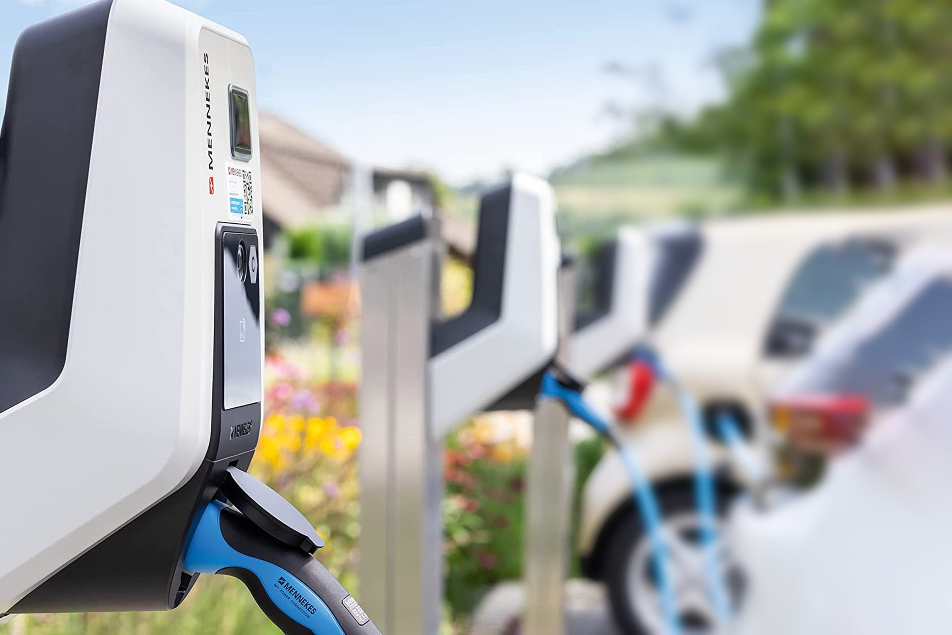 Die Ladestation Mennekes AMTRON® Professional von Mennekes, ausgestattet mit einem angeschlossenen Kabel und RFID-Autorisierung für sicheren Zugang, ist im Einsatz, während tagsüber in der Nähe ein weißes Auto lädt.