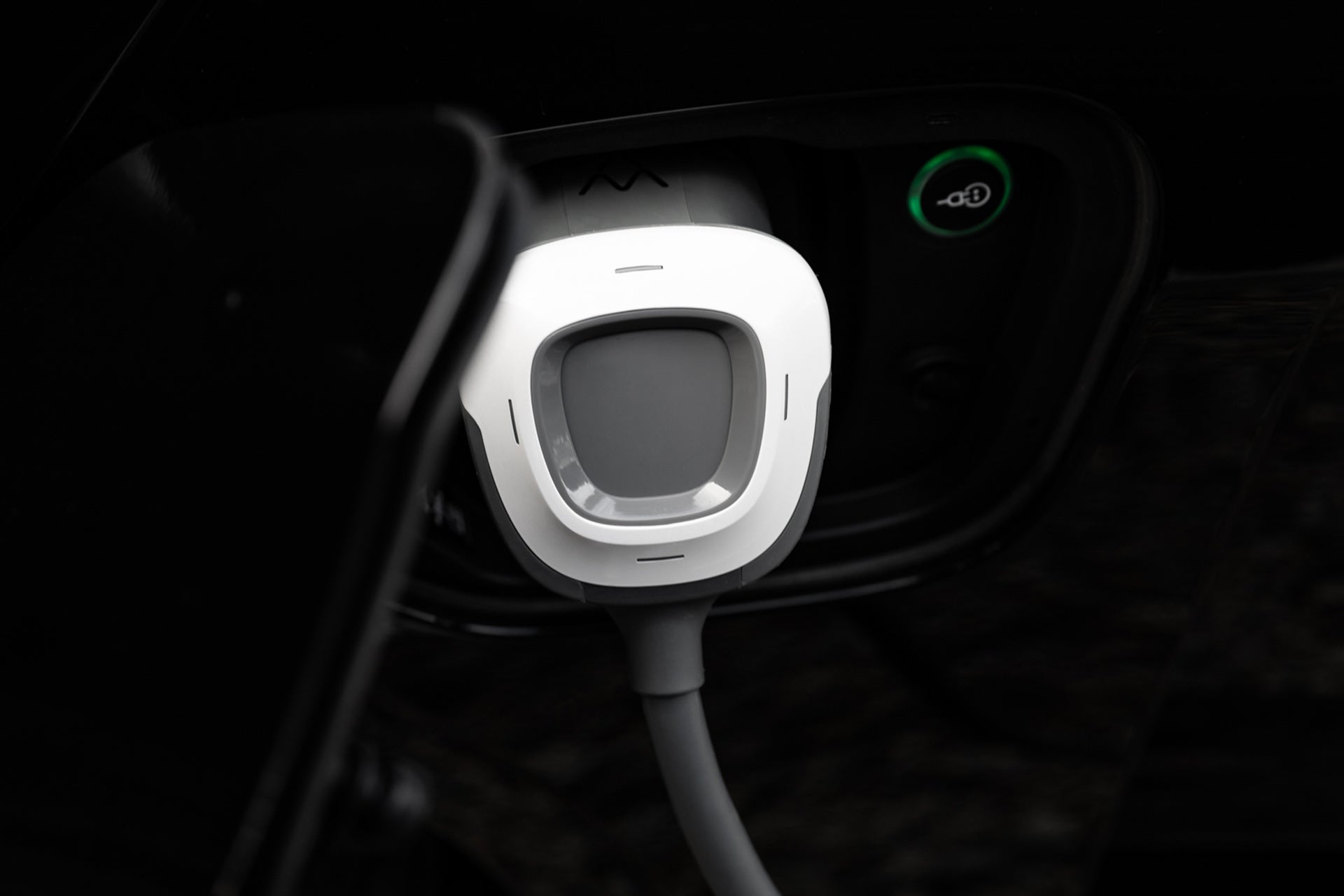 Nahaufnahme eines Charge Amps Aura-Ladesteckers von Charge Amps, der an ein Elektrofahrzeug angeschlossen ist, mit einer grünen Ladekontrollleuchte und zwei Ladepunkten im Hintergrund.