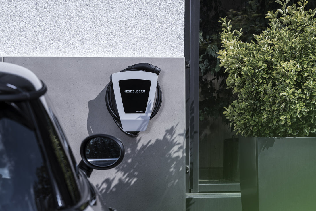 Eine Heidelberg AMPERFIED Wallbox connect.home Ladestation für Elektrofahrzeuge von Heidelberg AMPERFIED ist an einer Außenwand in der Nähe eines geparkten Autos und einer großen Topfpflanze montiert.
