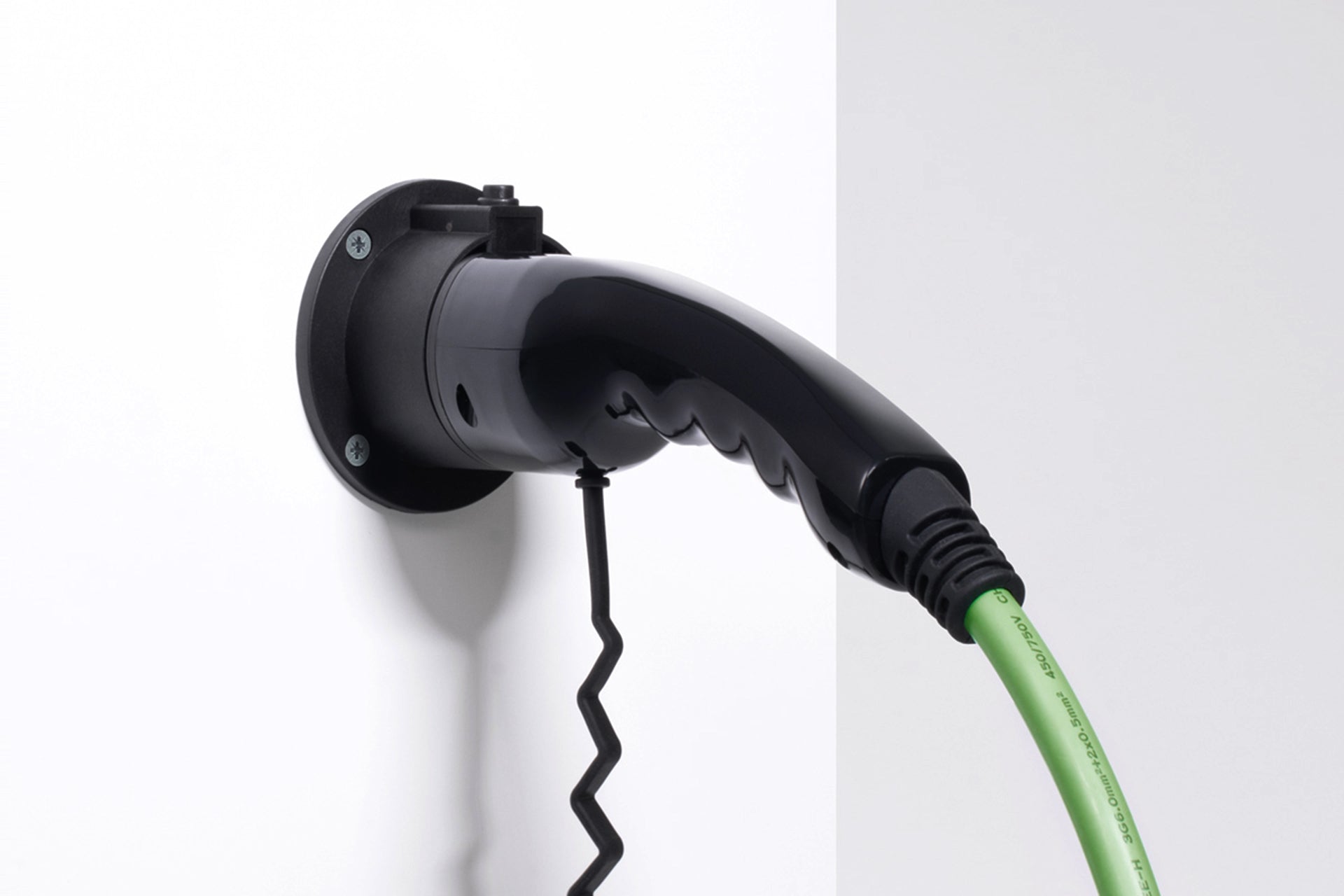 Die ELECTRIC-WAYS Steckerhalterung Typ 2 von ELECTRIC-WAYS befestigt einen schwarzen Typ 2 Ladestecker mit grünem Kabel sicher an einer weißen Wand und ermöglicht so eine einfache Installation.