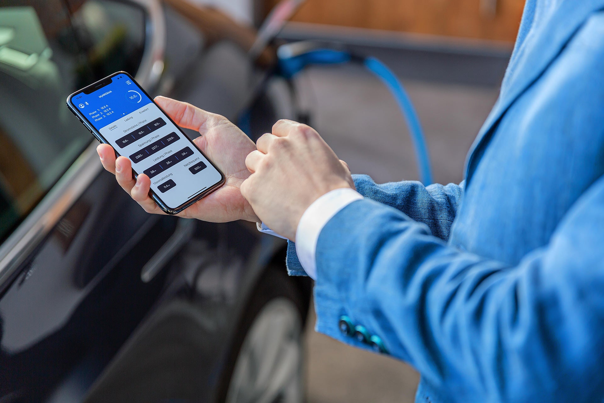 Eine Person im blauen Anzug überwacht per Smartphone-App den Ladevorgang eines Elektroautos mit 22 kW. Ein sichtbares Kabel und die mobile Ladeeinheit NRGkick Standard von NRGkick sorgen für eine zuverlässige Stromversorgung.