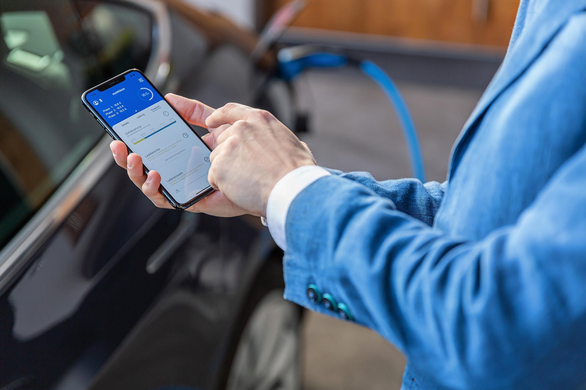 Eine Person im blauen Anzug nutzt eine Smartphone-App neben einem Elektroauto, das mit einem Typ-2-Stecker und einem NRGkick-Set von NRGkick lädt und bis zu 22 kW Ladeleistung bereitstellt.