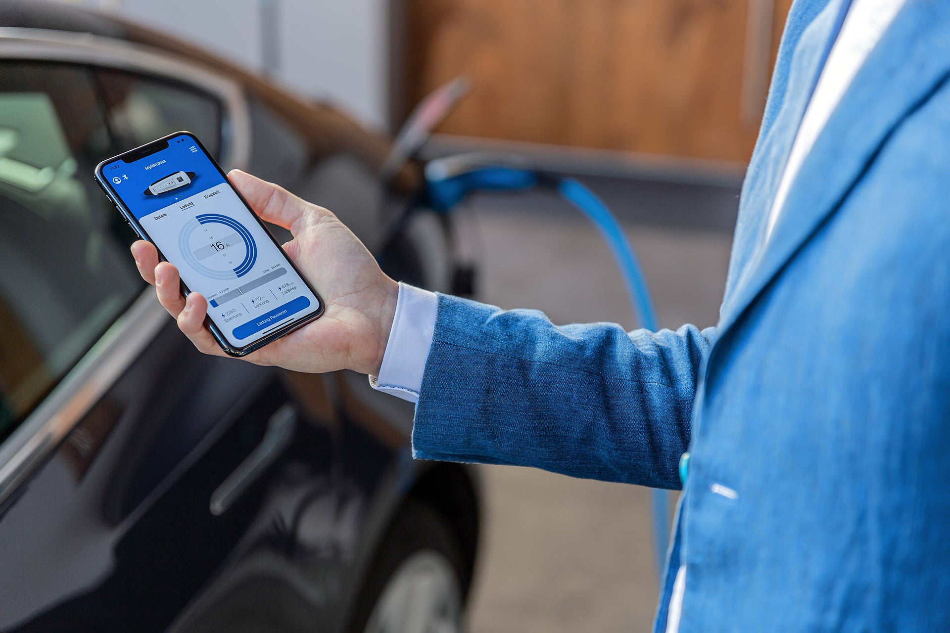 Eine Person in einem blauen Anzug hält ein Smartphone mit einer Autolade-App und steht neben einem Elektrofahrzeug, das mit der mobilen Ladeeinheit NRGkick Standard von NRGkick aufgeladen wird, die 22 kW Ladeleistung bietet.