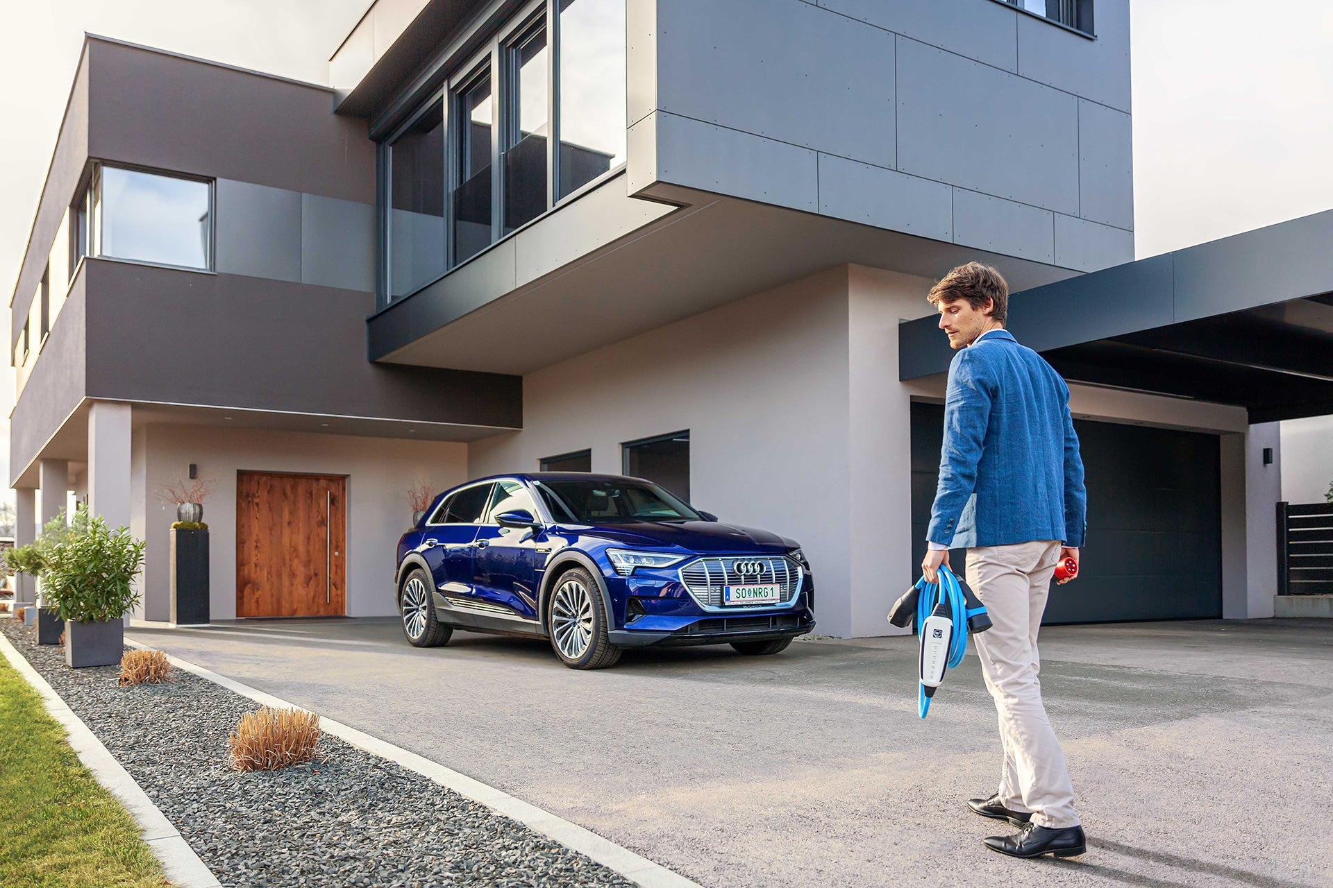 Ein Mann mit einem Typ-2-Stecker nähert sich einem blauen Elektroauto von Audi, das in einer Einfahrt geparkt ist, und bereitet sich darauf vor, es bequem mit seinem NRGkick-Set von NRGkick aufzuladen.