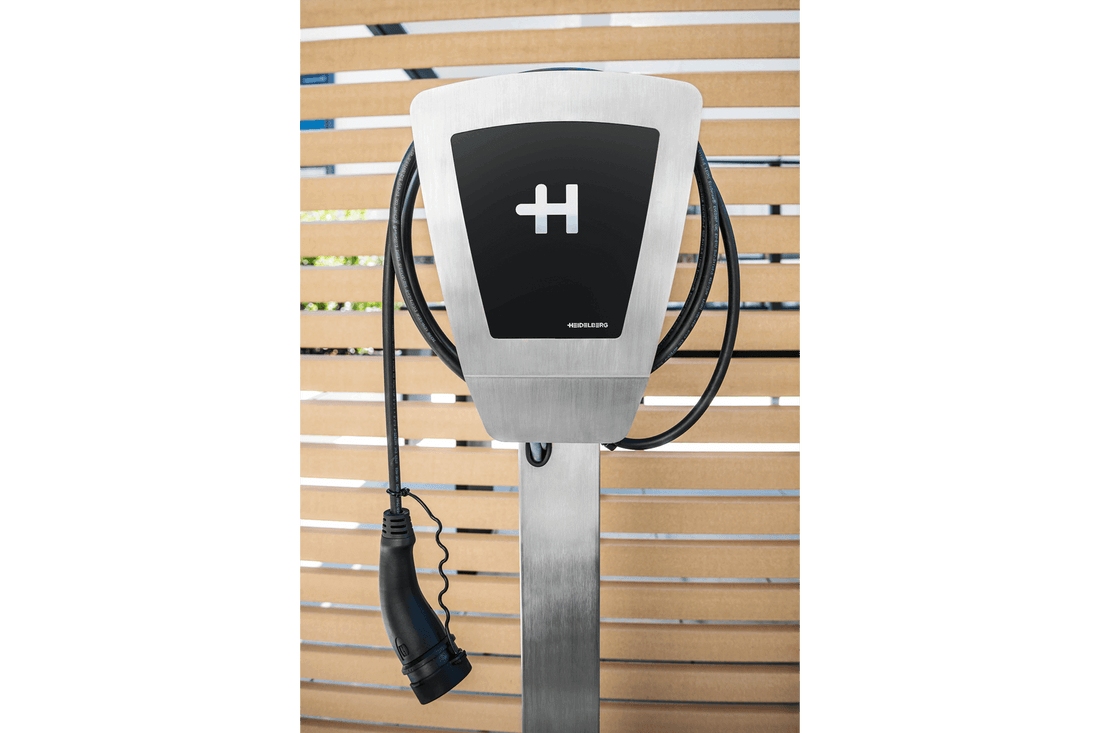 Vor Holzlatten wird die Heidelberg AMPERFIED Heidelberg Wallbox Home Eco gezeigt, eine Edelstahl-Ladestation für Elektrofahrzeuge mit Fehlerstromschutz, schwarzem Display und Spiralkabel.